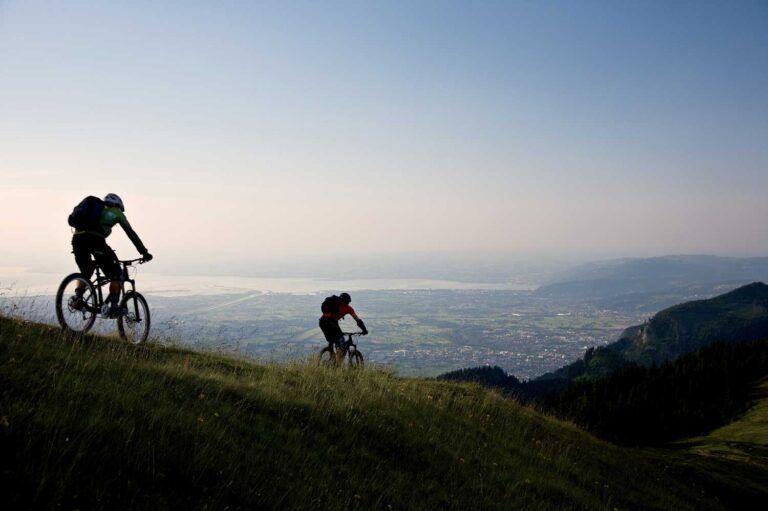 „Zwei Mountainbiker fahren auf einem Hügel bei der Hohe Kugel mit Blick auf Stadt und Bodensee im Hintergrund