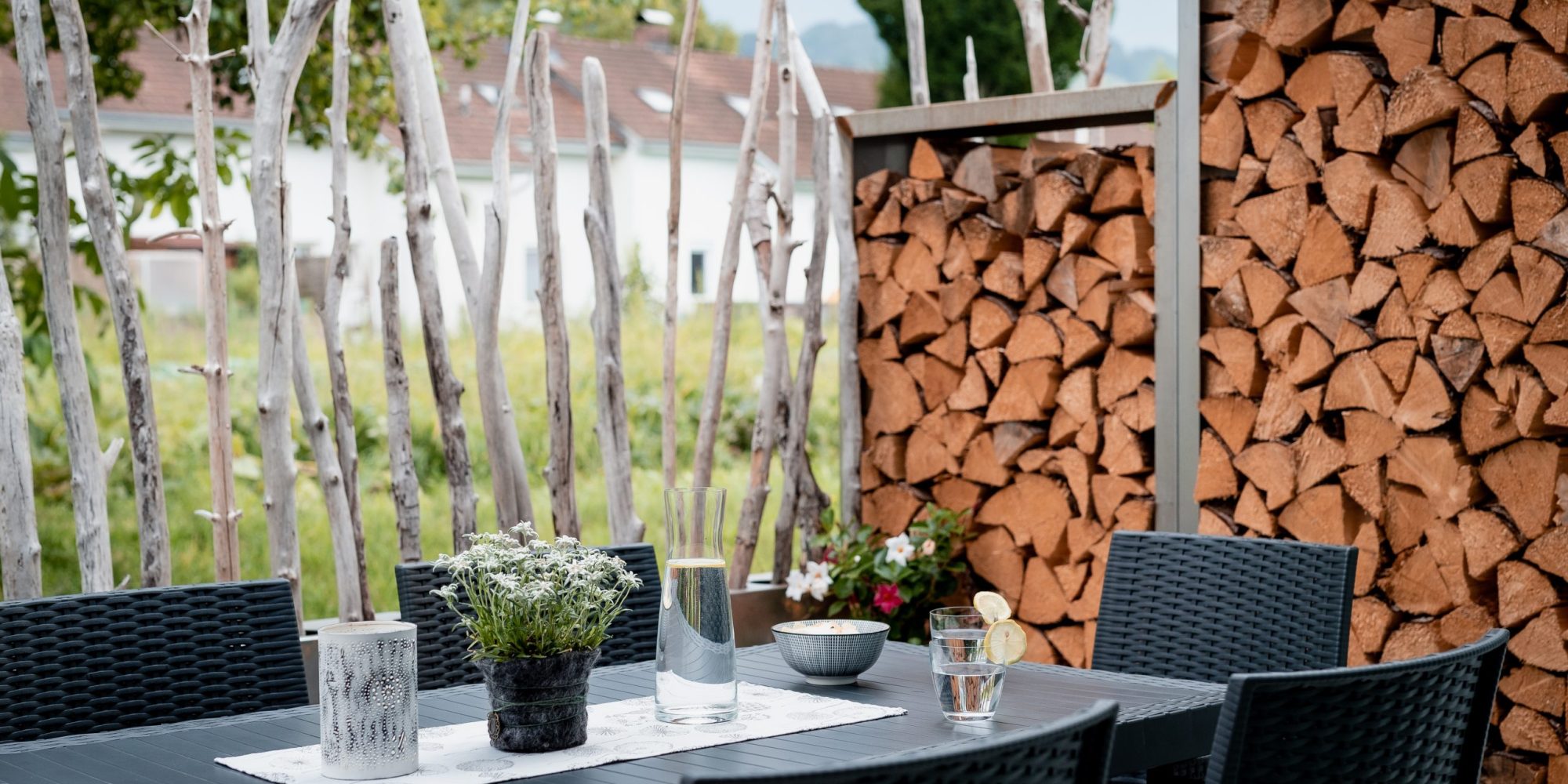 Ein Tisch mit schwarzen Stühlen im Freien, dekoriert mit Vase mit Blumen, Gläsern und Schale; im Hintergrund Holzstapel und Wand aus Ästen